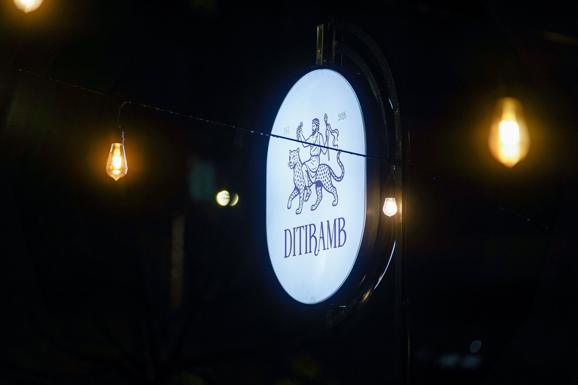 Illuminated oval sign for Ditiramb restaurant at night with warm-toned hanging lights
