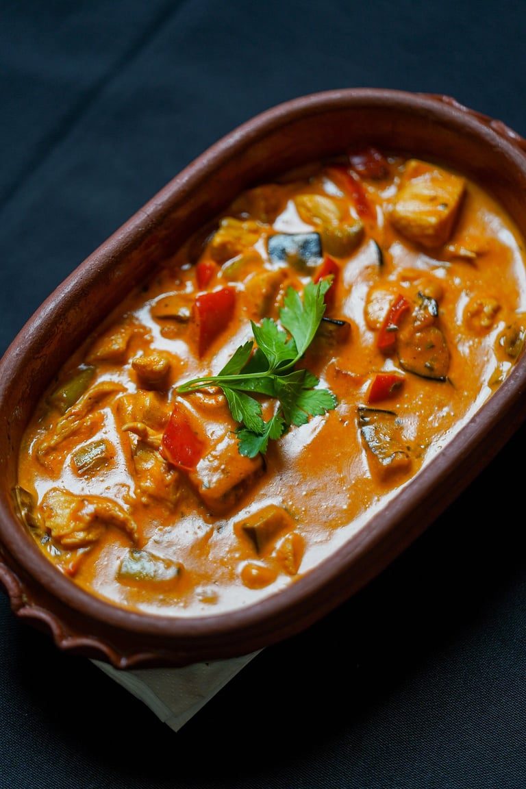 Creamy curry dish with chunks of meat and vegetables in a terracotta bowl, garnished with fresh parsley and red peppers