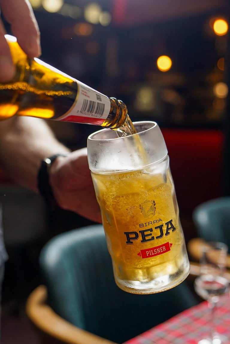 Birra Peja pilsner beer being poured from a bottle into a glass with bokeh lights in the background