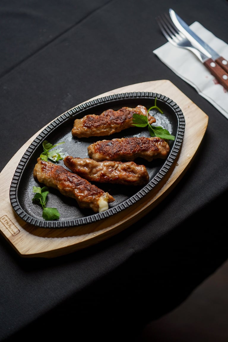 Four grilled sausages arranged on a wooden platter with fresh herbs, served with cutlery on a dark surface