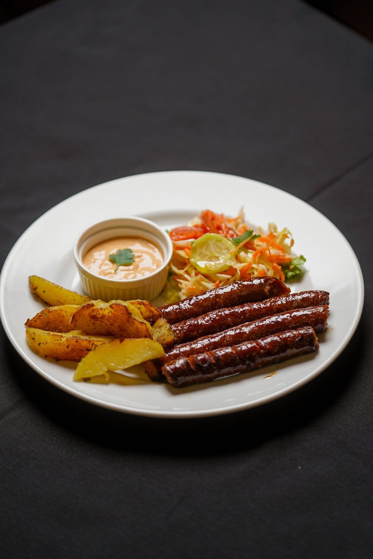 Plated meal with grilled sausages, crispy potato wedges, fresh vegetable salad, and creamy dipping sauce on a white plate against dark background